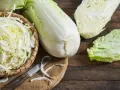 Frischer Chinakohl, bereit für leckere Chinakohl Rezepte. Ein ganzer Kohlkopf, geschnittene Blätter und fein gehackter Kohl in einem Korb liegen auf einem Holztisch.