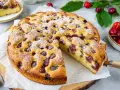 Ein saftiger Kirschkuchen, bestäubt mit Puderzucker, mit einem Stück herausgeschnitten. Frische Kirschen und Blätter liegen daneben.