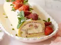 Erdbeer Biskuitrolle mit frischen Erdbeeren, Minze und Pistazien bestreut, auf einer weißen Servierplatte.