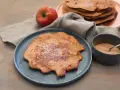 Ein Apfelpfannkuchen, bestreut mit Zucker und Zimt, liegt auf einem blauen Teller. Daneben ein roter Apfel und eine Schüssel mit Zimtzucker. Ein leckeres Apfelpfannkuchen Rezept zum Nachmachen.