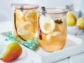 Zwei Gläser mit eingekochten Birnen, verfeinert mit Zimt und Sternanis. Ein frische Birne liegt daneben.