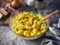 Schüssel mit warmem Kartoffelsalat, garniert mit Speck und Kräutern, daneben Zwiebeln und Speck.