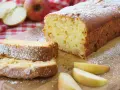 Ein saftiger, schneller Apfelkuchen mit Puderzucker bestäubt, daneben Apfelspalten und ganze Äpfel auf kariertem Tuch.