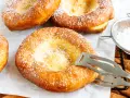 Frisch gebackene, ausgezogene Krapfen, bestäubt mit Puderzucker. Ein Kochlöffel hält einen der goldbraunen Krapfen.