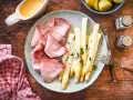 Spargel mit Schinken, garniert mit Petersilie, auf einem Teller mit Sauce Hollandaise und Kartoffeln daneben.