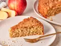 Ein saftiges Stück Apfel Haferflocken Kuchen liegt auf einem Teller, daneben ein ganzer Apfel und Haferflocken.