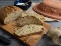 Frisch gebackenes Dinkelbrot mit Körnern, aufgeschnitten auf einem Holzbrett. Daneben ein Römertopf, eine Schüssel mit Joghurt und ein Messer.