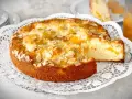 Ein goldbrauner Landfrauen Aprikosenkuchen, bestreut mit Mandelblättchen, wird auf einer zarten Tortenplatte serviert. Ein Stück wird gerade herausgehoben.