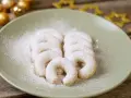 Vanillekipferl, bestäubt mit Puderzucker, liegen auf einem Teller. Goldene Kugeln und Sterne im Hintergrund deuten auf Weihnachten hin.