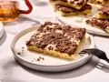 Ein Stück versunkener Streuselkuchen mit Schokostreuseln auf einem Teller, daneben eine Gabel und ein Glas Tee.