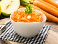 Frischer Karotten-Apfel-Salat in einer weißen Schüssel, garniert mit Petersilie. Im Hintergrund liegen ganze Karotten und ein halber Apfel.