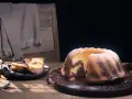 Ein fluffiger Marmorkuchen, bestäubt mit Puderzucker, liegt auf einem Holzbrett. Daneben zwei Stücke auf einem Teller.