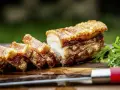 Knuspriger Schweinebauch, perfekt für ein köstliches schweinebauch rezept. Saftig, mit krosser Schwarte, auf einem Holzbrett mit Petersilie und einem Messer.