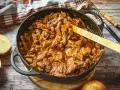 Herzhaftes Zwiebelfleisch-Rezept in Gusseisenpfanne, mit Zwiebeln und Fleischstücken in einer würzigen Sauce. Ein Holzlöffel mit der Aufschrift "Die Frau am Grill" liegt darin.
