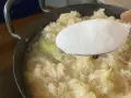 Zucker wird zu köchelndem Sauerkraut nach Omas Rezept gegeben. Ein Lorbeerblatt schwimmt im Topf.