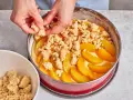 Hände verteilen Streusel auf einem Pfirsichkuchen in einer Springform.