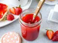 Ein Glas mit leuchtend roter Erdbeermarmelade, die mit einem Holzlöffel umgerührt wird. Daneben frische Erdbeeren und ein Stück Brot mit Marmelade – ein Genuss mit Pfiff!
