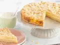 Ein saftiger Streuselkuchen mit Puddingfüllung, bestäubt mit Puderzucker. Daneben eine Tasse Cappuccino.