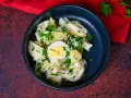 Ein cremiger Kartoffelsalat mit Mayo, garniert mit Petersilie und einem halben Ei, serviert in einer dunklen Schüssel.