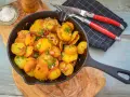 Bratkartoffeln mit Zwiebeln und Speck in einer Gusseisenpfanne, garniert mit Schnittlauch. Dazu grobes Salz und ein Messer-Gabel-Set.