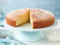 Ein **schneller saftiger Kuchen mit Joghurt**, bestäubt mit Puderzucker, auf einem hellblauen Tortenständer. Daneben eine Schale mit Sahne und Gabeln.