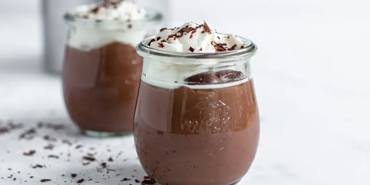 Zwei Gläser mit cremigem Schokopudding, gekrönt mit Schlagsahne und Schokostreuseln. Perfekt, um Schokopudding selber machen zu lernen!
