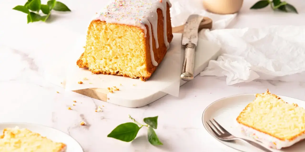 Saftiger zitronenkuchen kastenform mit Zuckerguss und bunten Streuseln, auf einem Marmorsockel, daneben ein Messer und ein Teller mit einem Stück Kuchen.