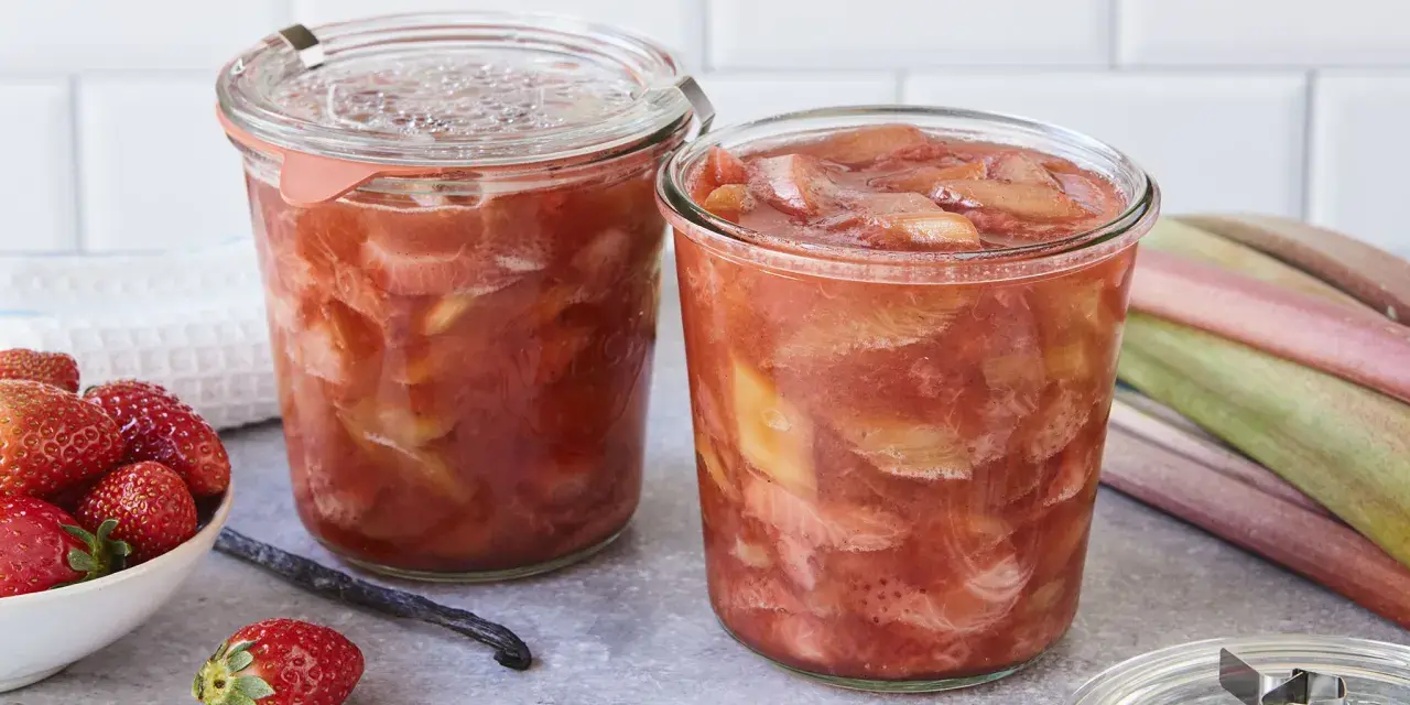 Zwei Gläser mit leckerem Rhabarberkompott, daneben frische Erdbeeren und Rhabarberstangen.