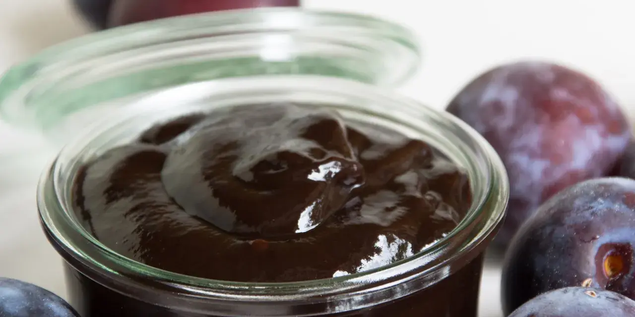 Ein Glas mit dunklem Pflaumenmus, umgeben von frischen Pflaumen. Perfekt für ein einfaches Pflaumenmus Rezept.