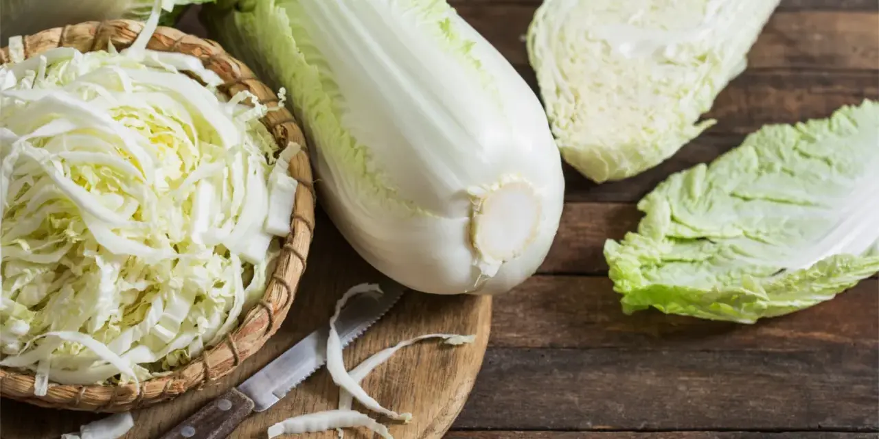 Frischer Chinakohl, bereit für leckere Chinakohl Rezepte. Ein ganzer Kohlkopf, geschnittene Blätter und fein gehackter Kohl in einem Korb liegen auf einem Holztisch.
