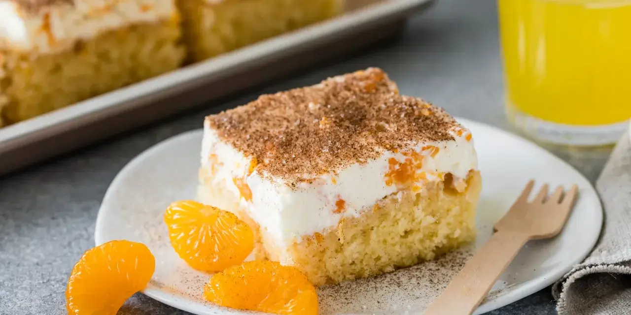 Ein saftiges Stück Fantakuchen-Rezept mit Mandarinen und Sahne, bestreut mit Zimt. Dazu ein Glas Fanta und frische Mandarinenstücke.