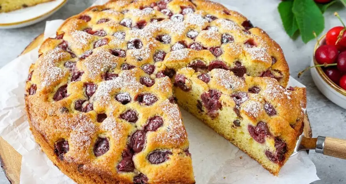 Ein saftiger Kirschkuchen, bestäubt mit Puderzucker, mit einem Stück herausgeschnitten. Frische Kirschen und Blätter liegen daneben.