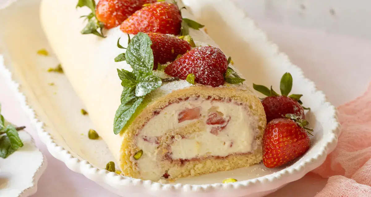 Erdbeer Biskuitrolle mit frischen Erdbeeren, Minze und Pistazien bestreut, auf einer weißen Servierplatte.