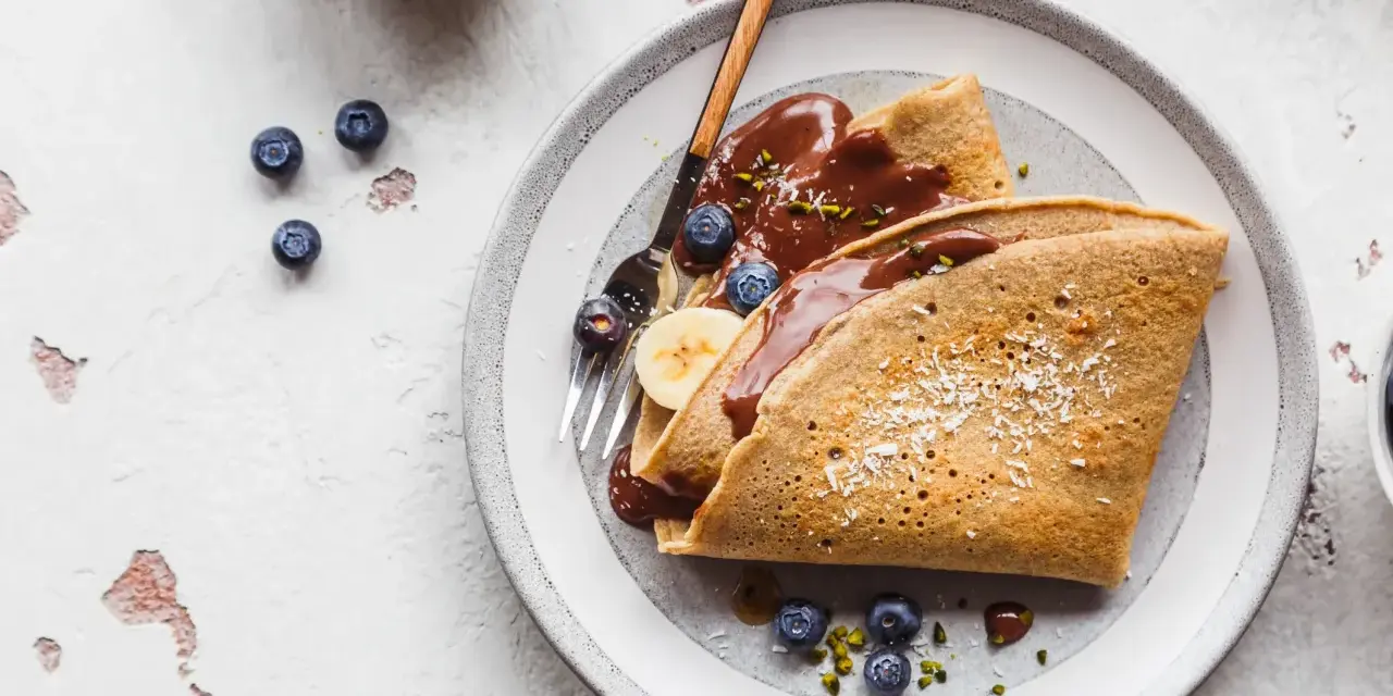 Buchweizen-Crêpes mit Schokoladensoße, Blaubeeren und Banane. Ein köstliches Beispiel für Buchweizen Rezepte, garniert mit Kokosraspeln und Pistazien.