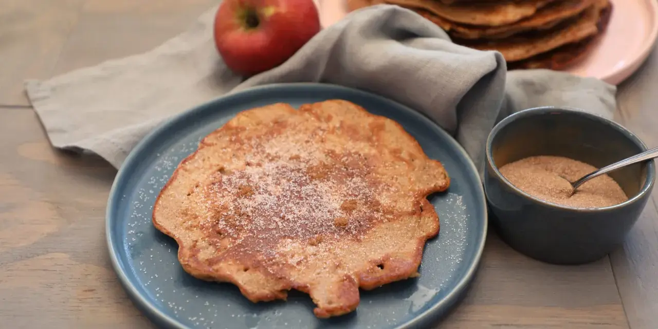 Ein Apfelpfannkuchen, bestreut mit Zucker und Zimt, liegt auf einem blauen Teller. Daneben ein roter Apfel und eine Schüssel mit Zimtzucker. Ein leckeres Apfelpfannkuchen Rezept zum Nachmachen.