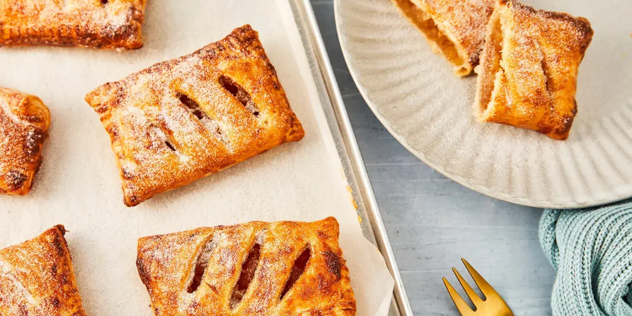 Goldbraune Apfeltaschen, bestäubt mit Puderzucker, warten auf dem Backblech und auf einem Teller. Ein köstliches Apfeltaschen Rezept zum Nachbacken.