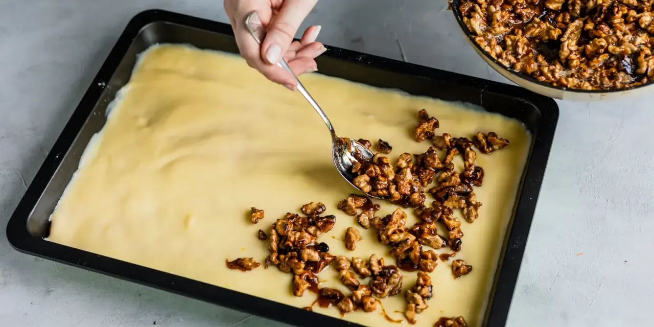 Eine Hand verteilt karamellisierte Walnüsse auf einem hellgelben Teig in einer Backform. Bald gibt es einen "friss dich dumm Kuchen"!