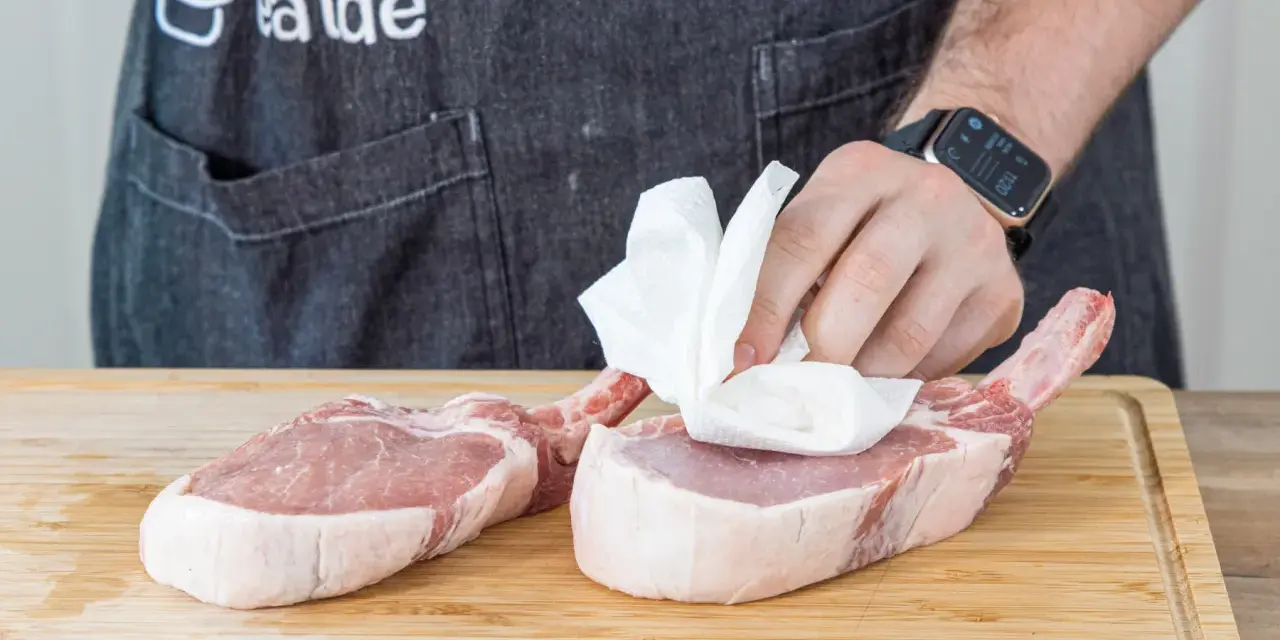 Vorbereitung für ein saftiges Kotelett Rezept: Frische Schweinekoteletts werden auf einem Holzbrett mit Küchenpapier abgetupft.