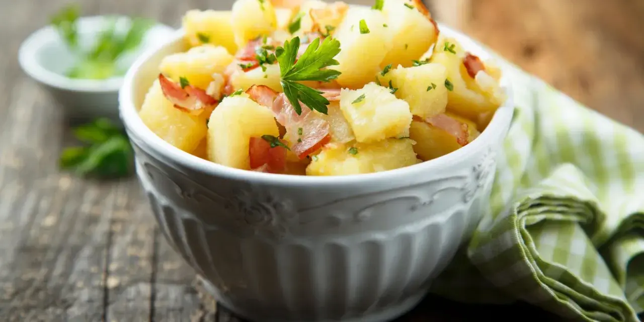 Ein einfacher Kartoffelsalat mit Speck und Petersilie in einer Schüssel, serviert auf einem Holztisch.