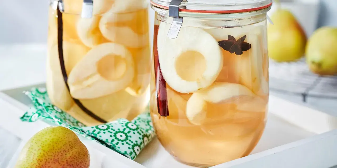 Zwei Gläser mit eingekochten Birnen, verfeinert mit Zimt und Sternanis. Ein frische Birne liegt daneben.