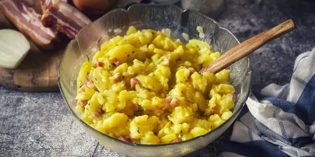Schüssel mit warmem Kartoffelsalat, garniert mit Speck und Kräutern, daneben Zwiebeln und Speck.