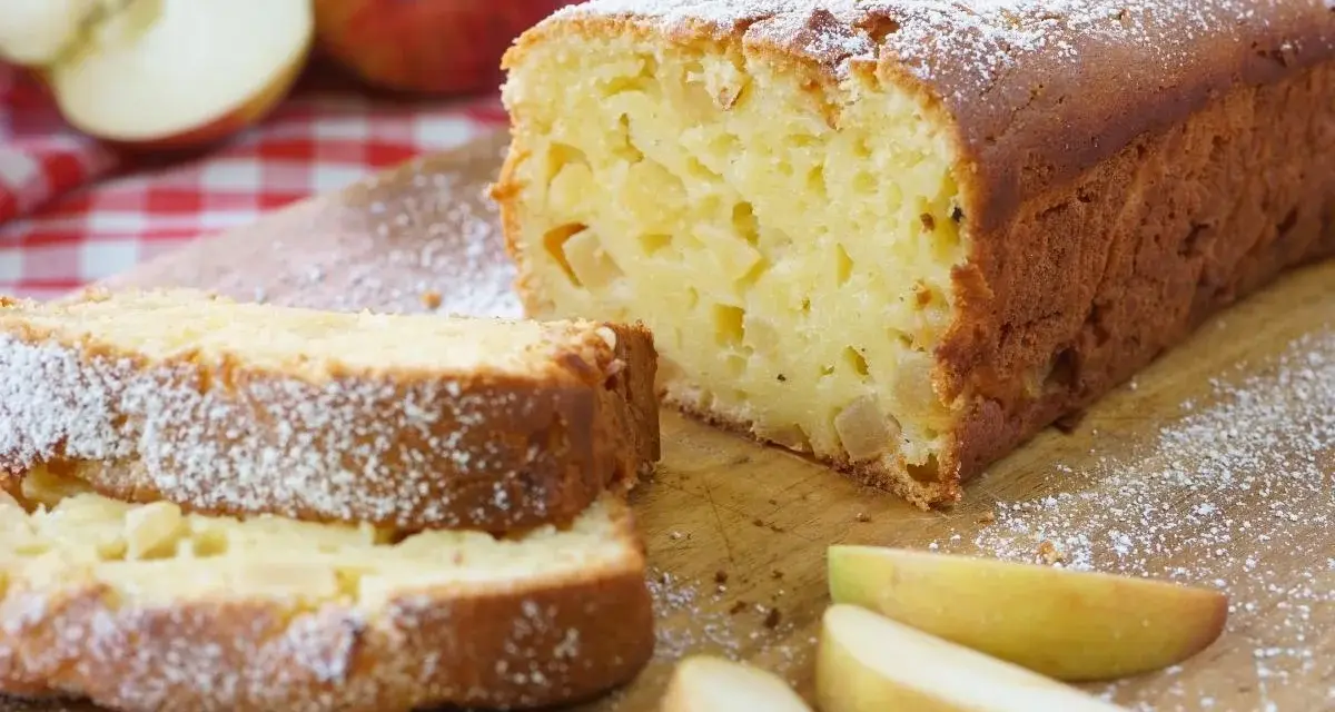 Ein saftiger, schneller Apfelkuchen mit Puderzucker bestäubt, daneben Apfelspalten und ganze Äpfel auf kariertem Tuch.