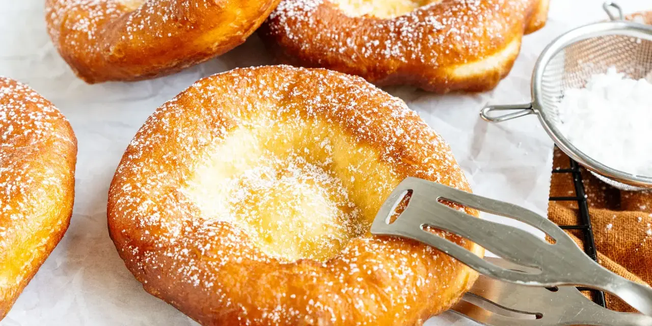 Frisch gebackene, ausgezogene Krapfen, bestäubt mit Puderzucker. Ein Kochlöffel hält einen der goldbraunen Krapfen.