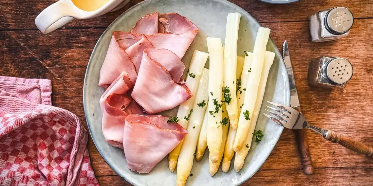 Spargel mit Schinken, garniert mit Petersilie, auf einem Teller mit Sauce Hollandaise und Kartoffeln daneben.