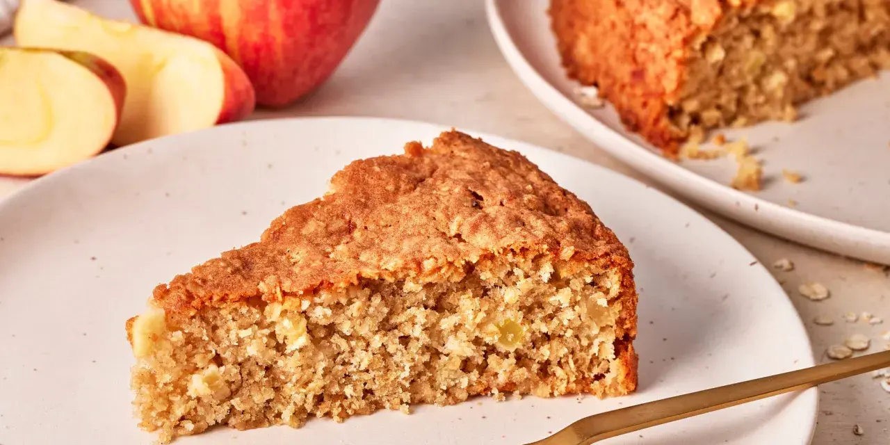 Ein saftiges Stück Apfel Haferflocken Kuchen liegt auf einem Teller, daneben ein ganzer Apfel und Haferflocken.