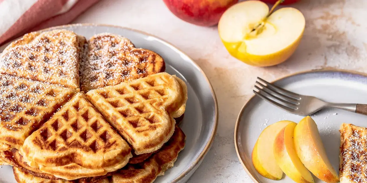 Herzförmige Waffeln mit Puderzucker, daneben frische Äpfel und Apfelspalten. Ein Genuss für alle Sinne!