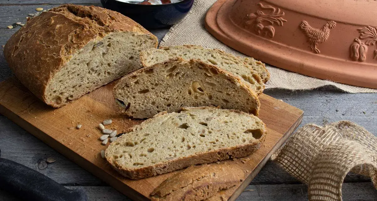 Frisch gebackenes Dinkelbrot mit Körnern, aufgeschnitten auf einem Holzbrett. Daneben ein Römertopf, eine Schüssel mit Joghurt und ein Messer.