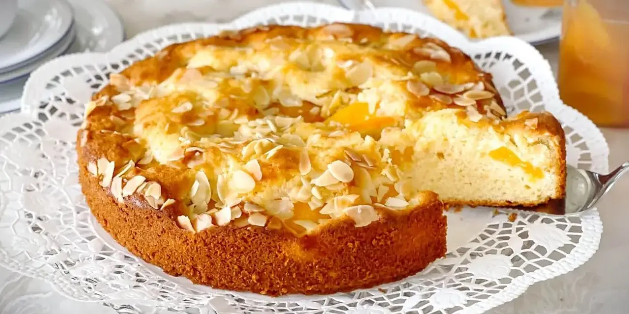 Ein goldbrauner Landfrauen Aprikosenkuchen, bestreut mit Mandelblättchen, wird auf einer zarten Tortenplatte serviert. Ein Stück wird gerade herausgehoben.
