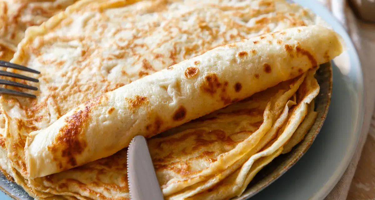 Ein Stapel goldbrauner Pfannkuchen, einer davon aufgerollt, mit Messer und Gabel. Perfekt für ein leckeres Pfannkuchen Rezept!