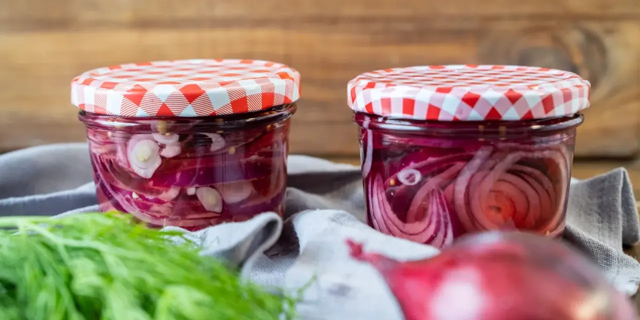 Zwei Gläser mit eingelegten Zwiebeln, rot und weiß, stehen auf einem grauen Tuch. Frischer Dill und eine rote Zwiebel liegen davor.
